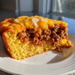 A thick slice of sloppy joe cornbread casserole topped with melted cheddar cheese on a white plate.