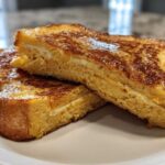 Two thick slices of golden brown sourdough french toast stacked slightly on a white plate.