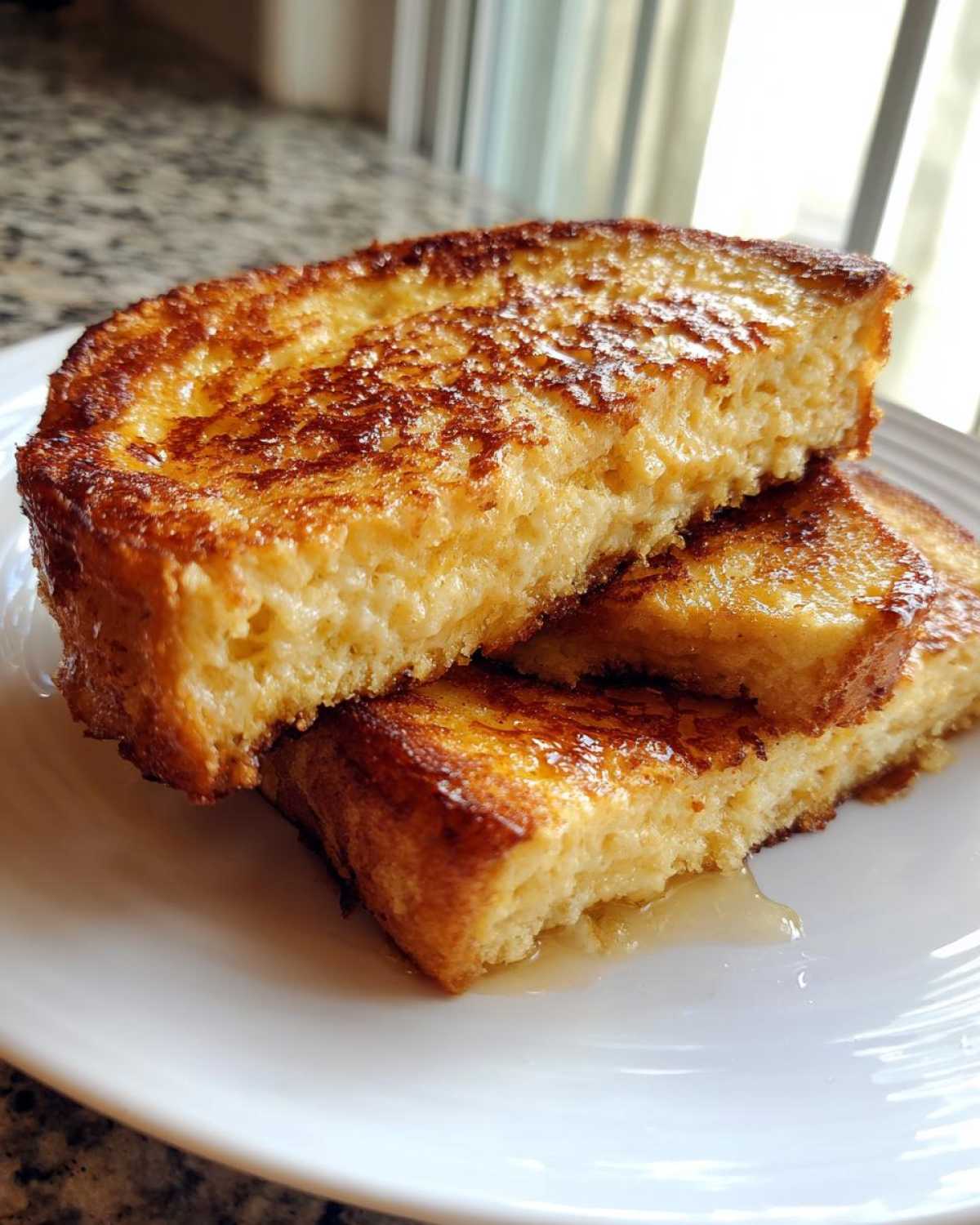 Close-up of thick, golden-brown sourdough french toast stacked on a white plate with syrup pooling underneath.
