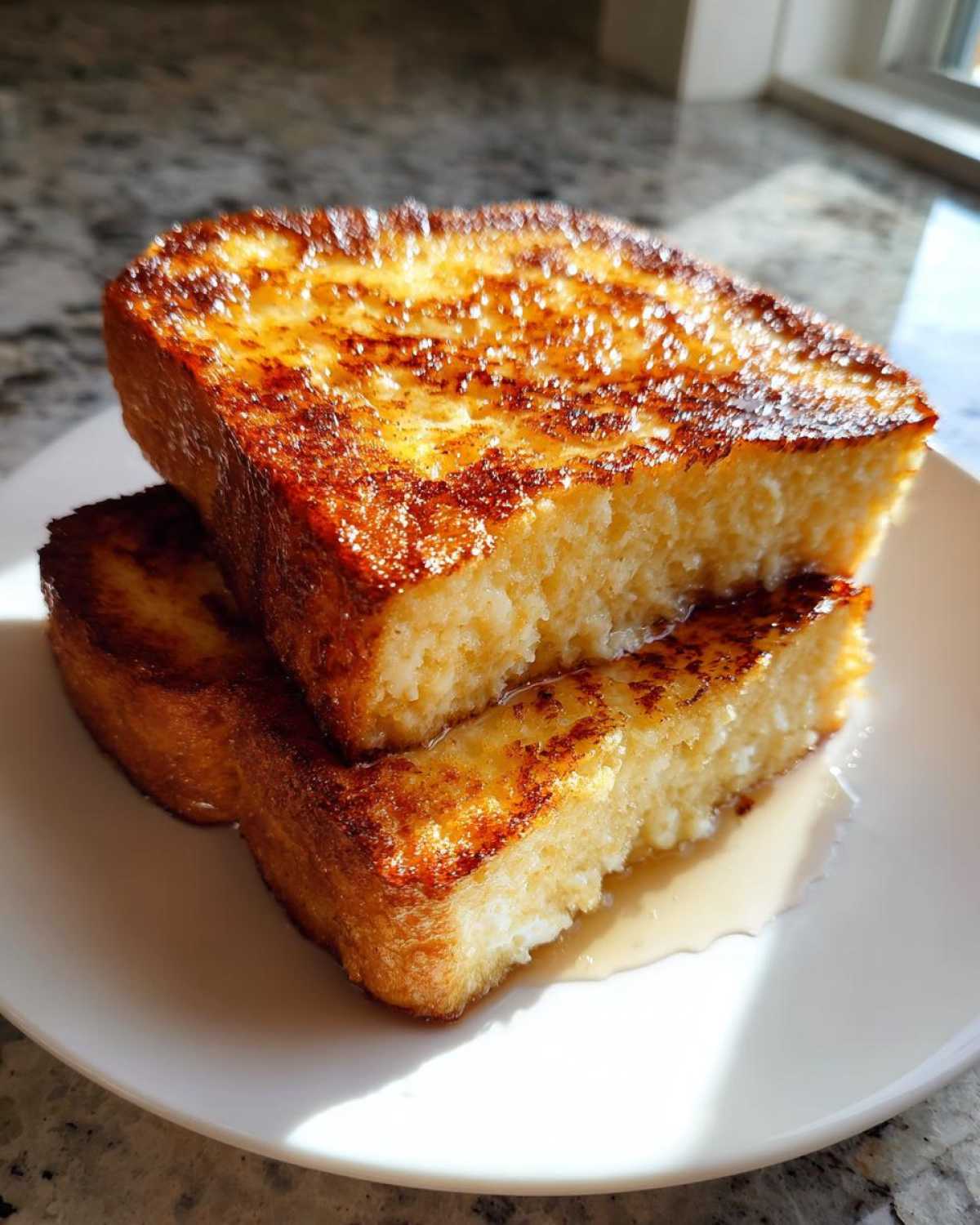 Two thick slices of golden brown sourdough french toast stacked on a white plate with syrup pooling underneath.
