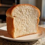 A thick, freshly baked slice of soft sourdough sandwich bread standing upright on a white plate.