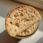 Overhead view of a slice of bread with an open, airy crumb structure resting on top of a jar containing active sourdough starter.