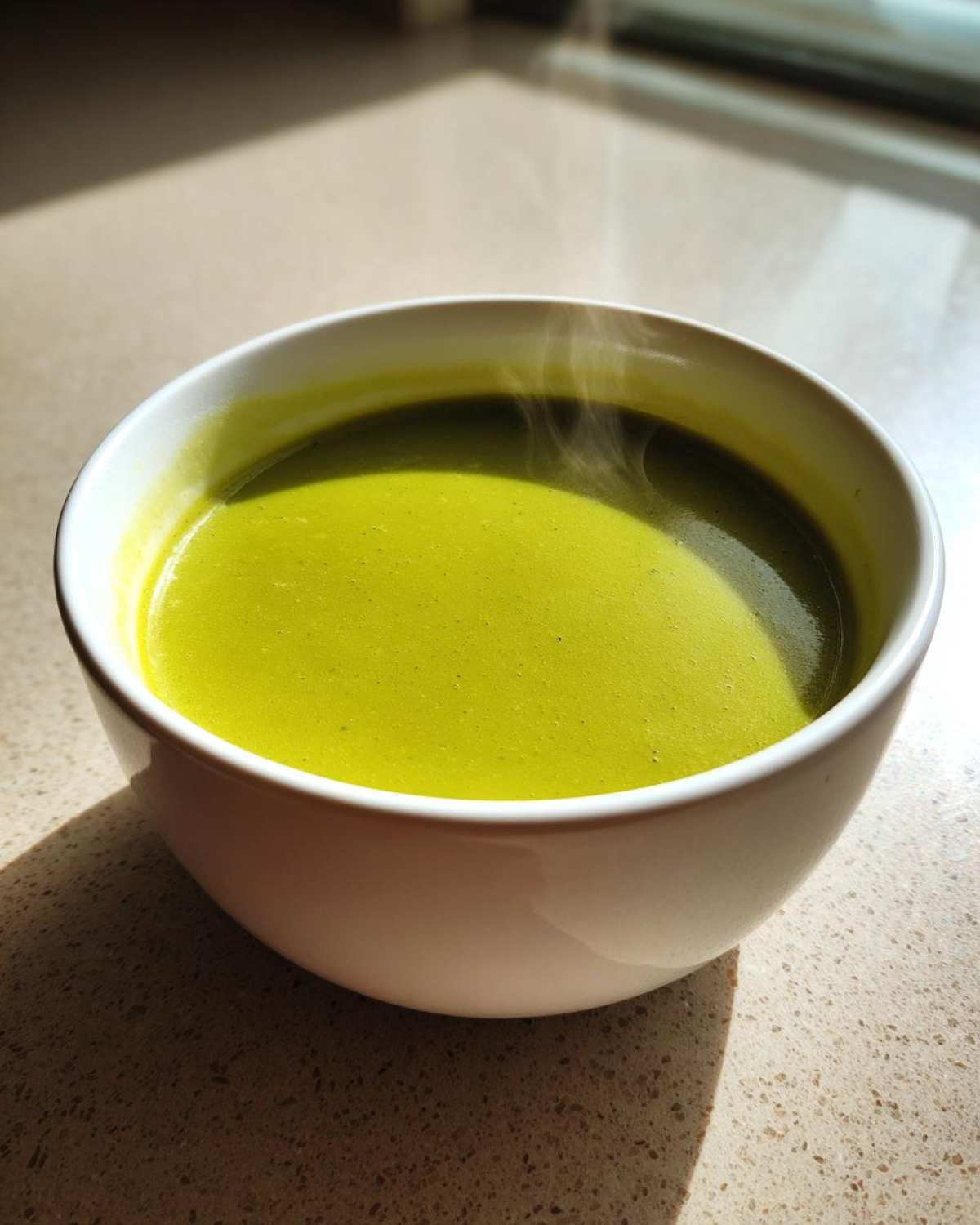 Close-up of a white bowl filled with hot, bright green zucchini soup, showing steam rising.