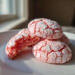 Close-up of pink, cracked strawberry cake mix cookies dusted heavily with powdered sugar on a white plate.