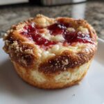Close-up of a single, freshly baked strawberry cheesecake cookie with a golden-brown crumb topping and bright red strawberry jam center.