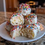 A stack of white chocolate-dipped sugar cookie truffles covered in rainbow sprinkles, one is cut open.