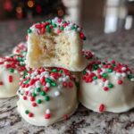 A stack of white chocolate coated sugar cookie truffles topped with red, green, and white sprinkles, one cut open.