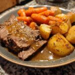 Close-up of a plate featuring tender pot roast covered in rich brown gravy alongside roasted potatoes and glazed carrots.