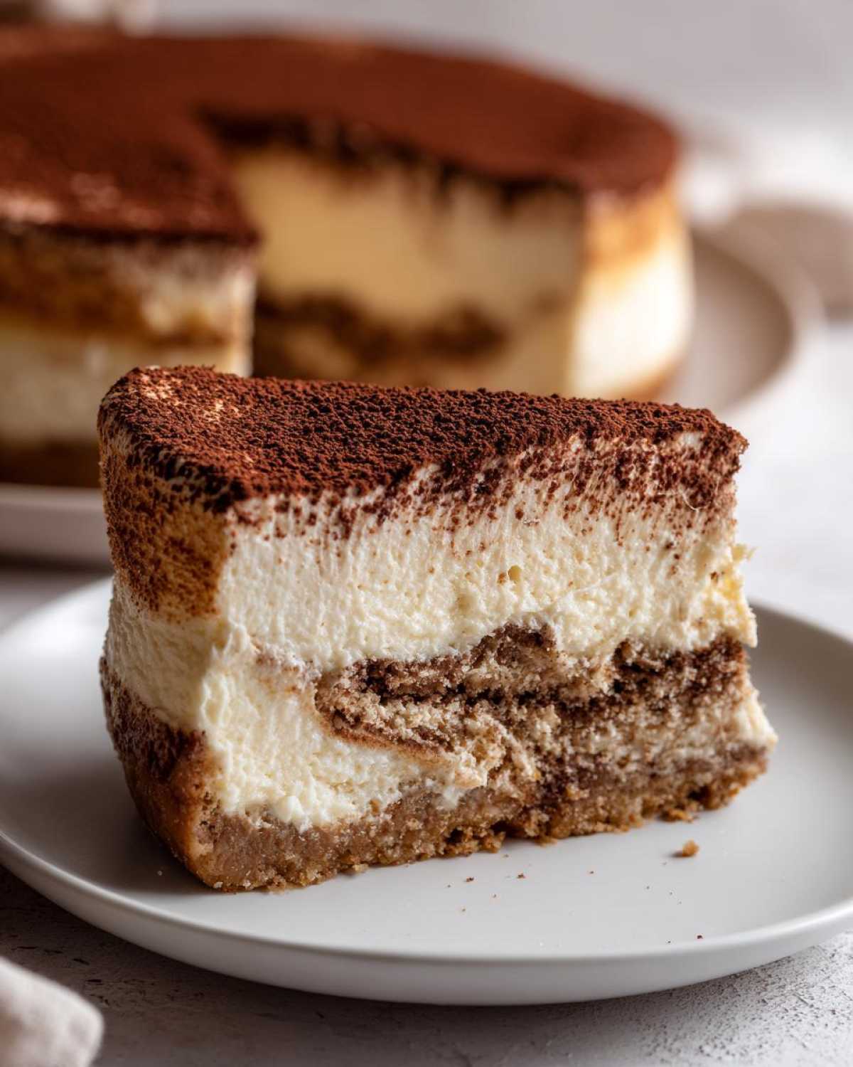 A thick slice of tiramisu cheesecake with creamy layers and a cocoa powder topping, sitting on a white plate.