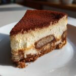 A perfect slice of tiramisu cheesecake showing layers of cream, coffee-soaked ladyfingers, and a heavy dusting of cocoa powder.
