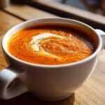 Close-up of rich, orange tomato basil soup swirled with cream and sprinkled with herbs in a white mug.