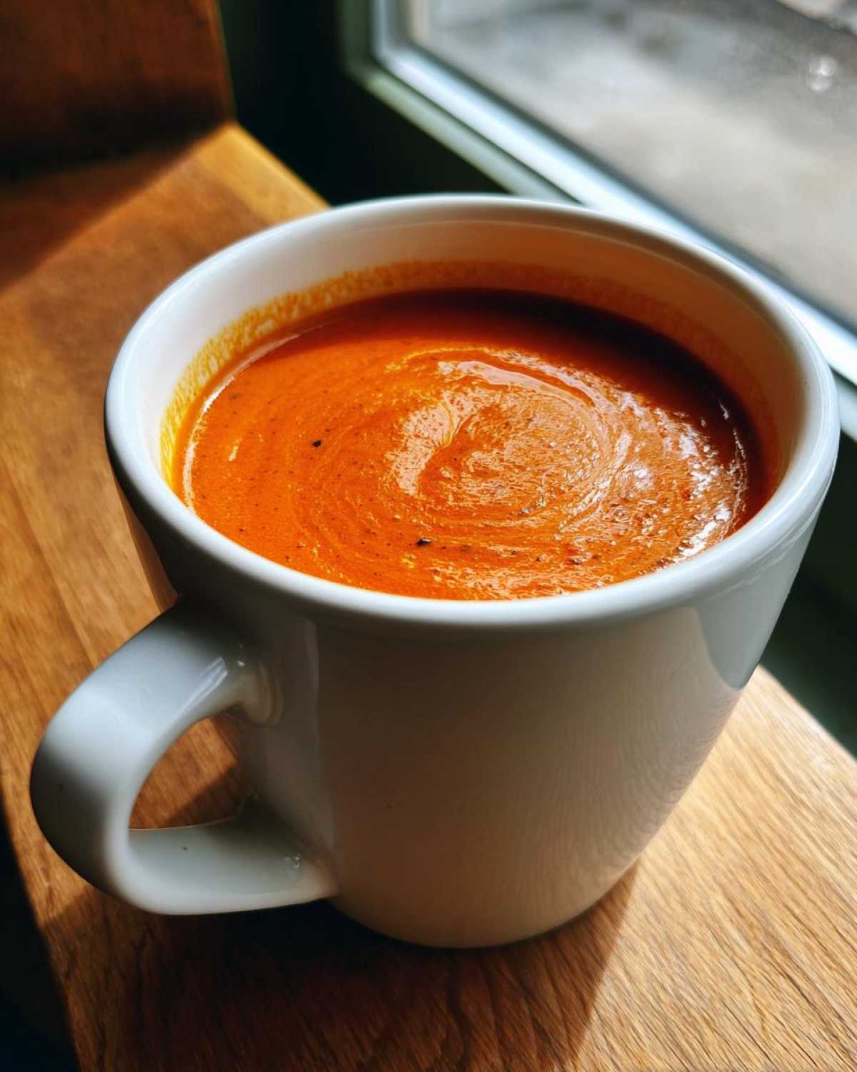 A close-up of vibrant orange tomato basil soup served in a white mug, sitting on a wooden surface near a window.