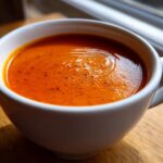 Close-up of rich, orange-red tomato basil soup sprinkled with pepper in a white mug.