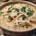 Close-up of a bowl of creamy white chicken chili with shredded chicken, white beans, topped with shredded cheese and fresh cilantro.