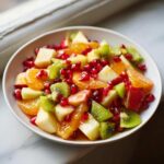 Close-up of a bright winter fruit salad featuring kiwi, oranges, apples, and pomegranate seeds in a white bowl.
