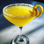 A vibrant yellow, icy Lemon Drop cocktail served in a sugar-rimmed coupe glass with a citrus twist garnish.