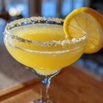 Close-up of a bright yellow lemon drop cocktail served in a coupe glass with a sparkling sugared rim and lemon wheel garnish.