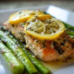 Close-up of a baked salmon recipe fillet topped with lemon slices and herbs, served alongside bright green asparagus.