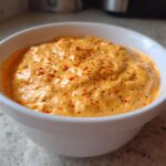 Close-up of a creamy, orange-hued beer cheese dip, topped with visible black pepper and red pepper flakes.