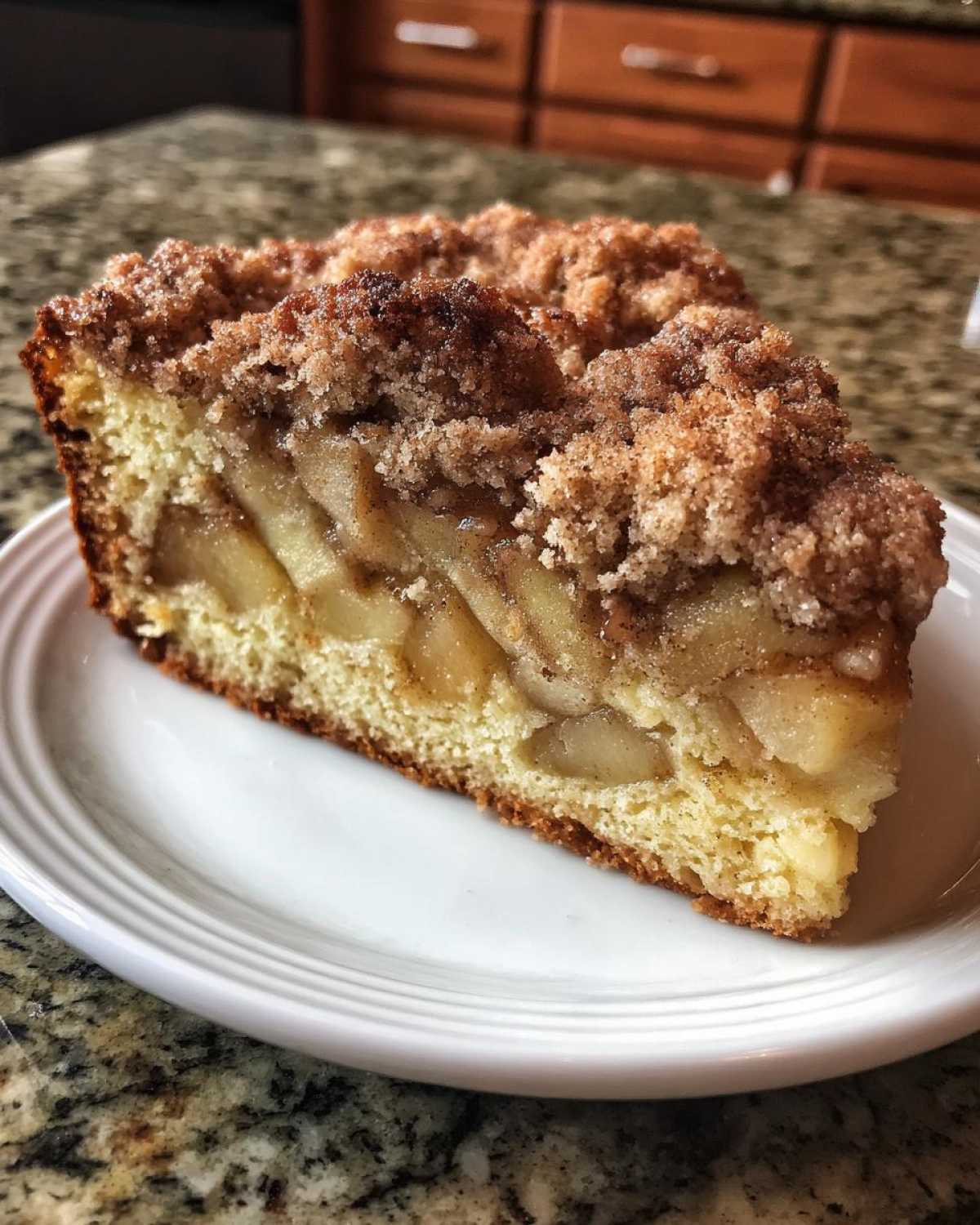A close-up of a thick slice of apple crumb cake showing layers of moist cake, spiced apples, and a crunchy cinnamon streusel topping.