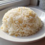 A perfectly molded mound of fluffy, white butter rice served on a bright white plate near a window.