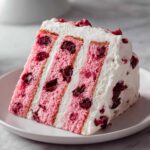 A tall slice of pink layered cherry cake filled and frosted with white whipped cream and whole cherries.