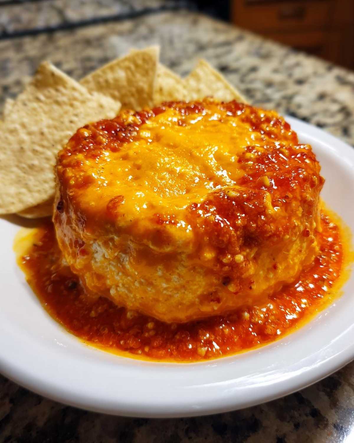 A round serving of hot chili cheese dip topped with melted orange cheese, surrounded by sauce, served with tortilla chips.