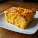 A square slice of moist, golden corn casserole packed with whole corn kernels, served on a white plate.
