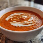 Close-up of a white bowl filled with vibrant orange tomato bisque, topped with a swirl of cream and herbs.