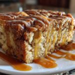 A close-up of a thick slice of apple bread pudding topped with caramel and vanilla icing.