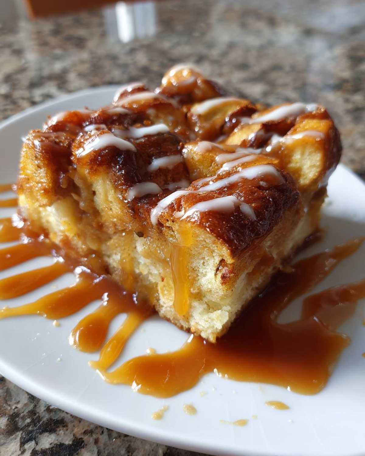 A square serving of apple bread pudding drizzled heavily with caramel sauce and white icing.