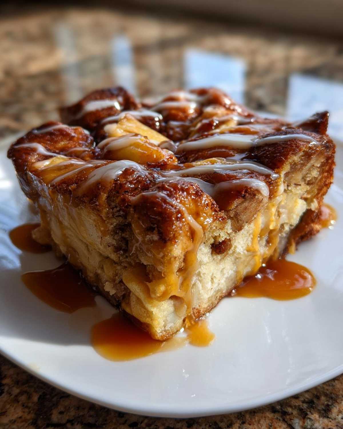 A close-up of a square serving of apple bread pudding, drizzled with white icing and rich caramel sauce.