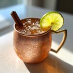 A condensation-covered copper mug filled with an apple cider moscow mule, garnished with a lime wheel and cinnamon stick.