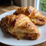 Two golden brown baked ham and cheese croissants with melted cheese oozing out, served on a white plate.