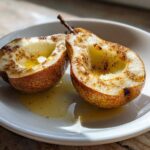 Two halves of warm baked pears sprinkled with cinnamon resting in syrup on a white plate.