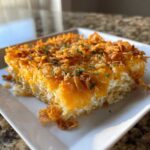 A close-up of a square serving of cheesy potato casserole topped with melted cheddar and crispy fried onions.