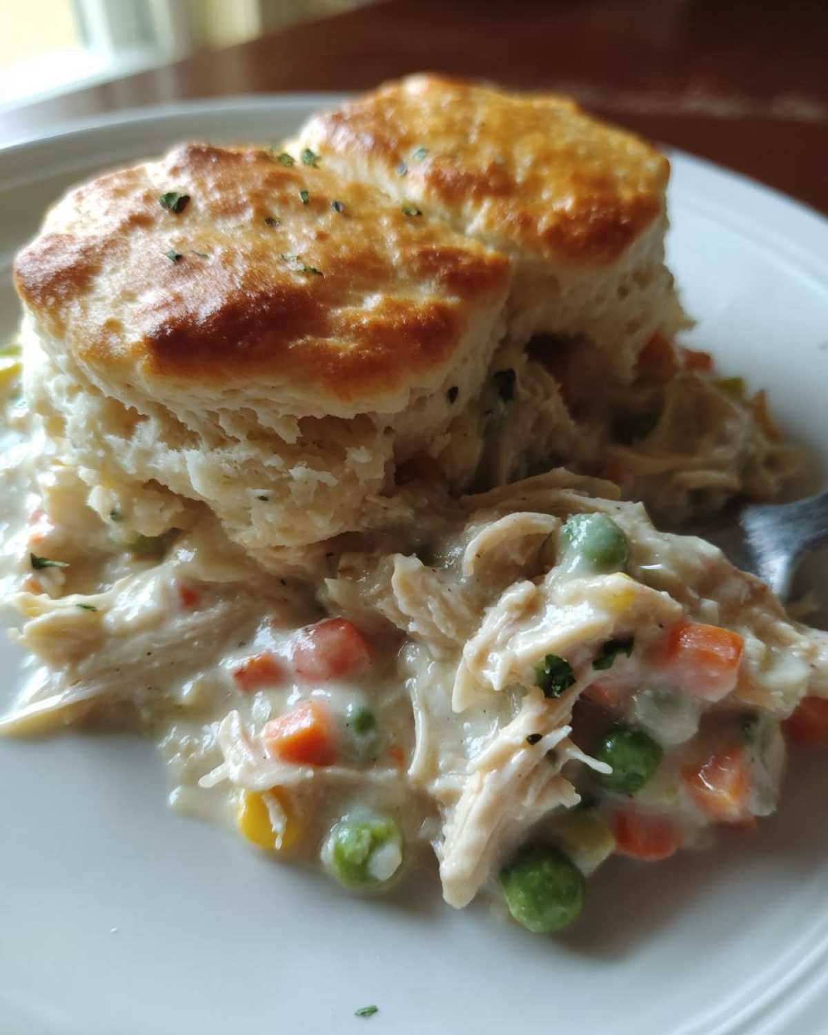 A close-up of a serving of creamy chicken pot pie casserole topped with golden biscuits, showing shredded chicken and mixed vegetables.