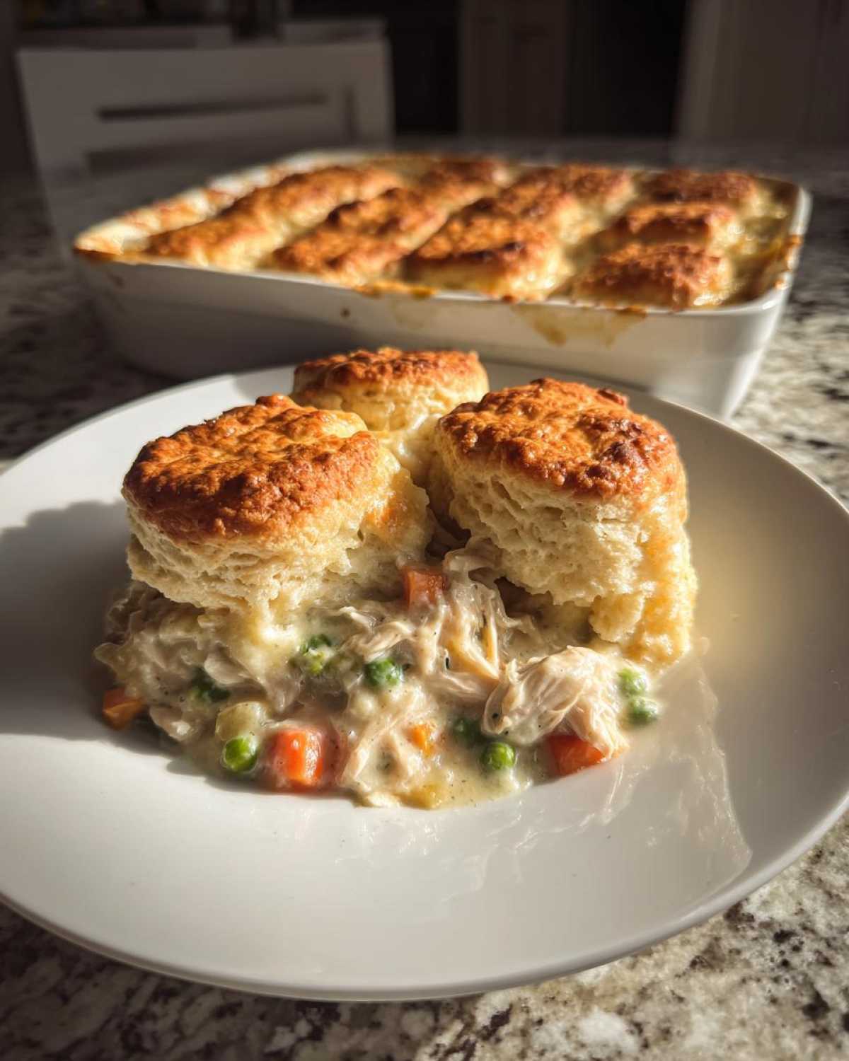 A serving of creamy chicken pot pie casserole topped with three golden biscuits, with the baking dish in the background.