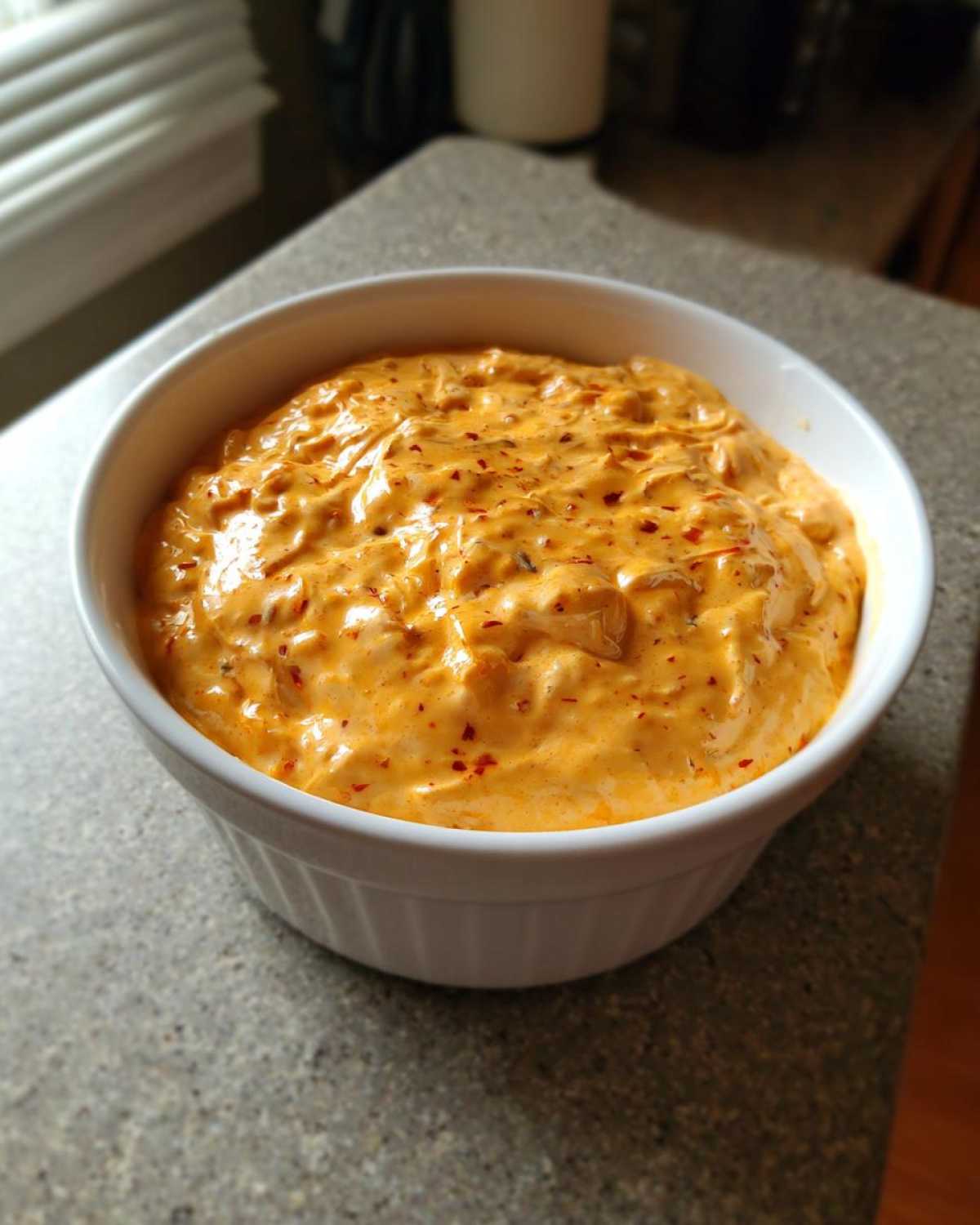 A white bowl filled with thick, creamy orange beer cheese dip, speckled with red pepper flakes, sitting on a counter.