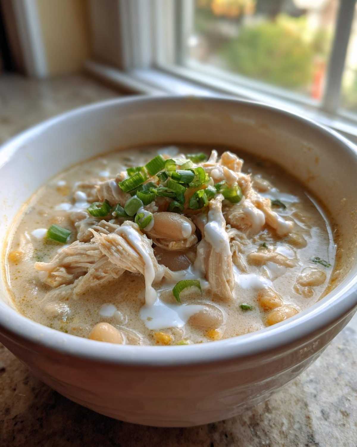 A close-up of a white bowl filled with creamy chicken chili, topped with shredded chicken, white beans, and green onions.