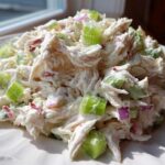 A mound of creamy crab salad featuring shredded crab meat, bright green celery chunks, and specks of red onion on a white plate.