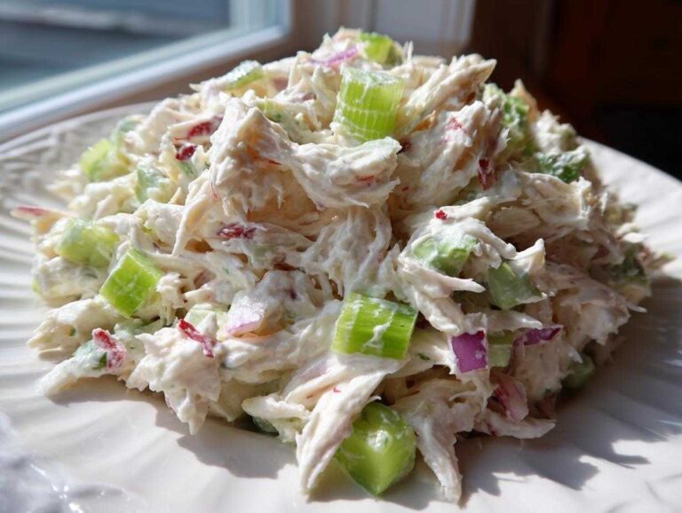 A mound of creamy crab salad featuring shredded crab meat, bright green celery chunks, and specks of red onion on a white plate.