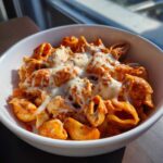 Close-up of a white bowl filled with creamy enchilada pasta, chicken pieces, and melted white cheese on top.