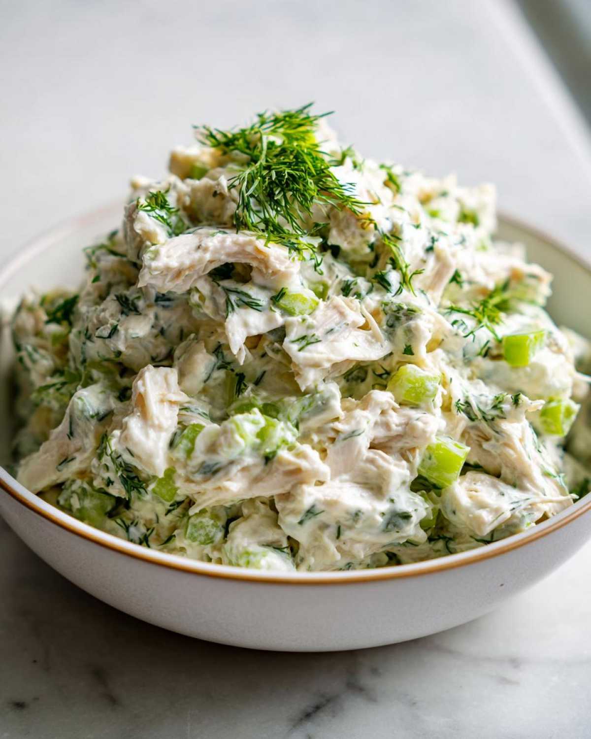 A close-up of a bowl filled with creamy greek yogurt chicken salad mixed with celery and topped with fresh dill.