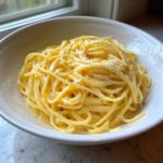 A close-up of a generous serving of creamy lemon pasta tossed in a white bowl, topped with lemon zest.