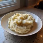A generous serving of creamy, fluffy mashed potatoes in a white bowl on a speckled countertop.
