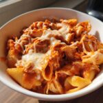 Close-up of creamy enchilada pasta with melted cheese in a white bowl, featuring shell pasta and shredded chicken.
