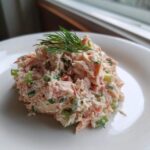 A mound of creamy salmon salad mixed with celery and herbs, garnished with fresh dill, served on a white plate.