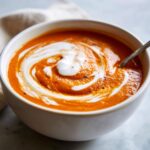 Close-up of a rich, orange tomato bisque topped with a swirl of cream and black pepper.
