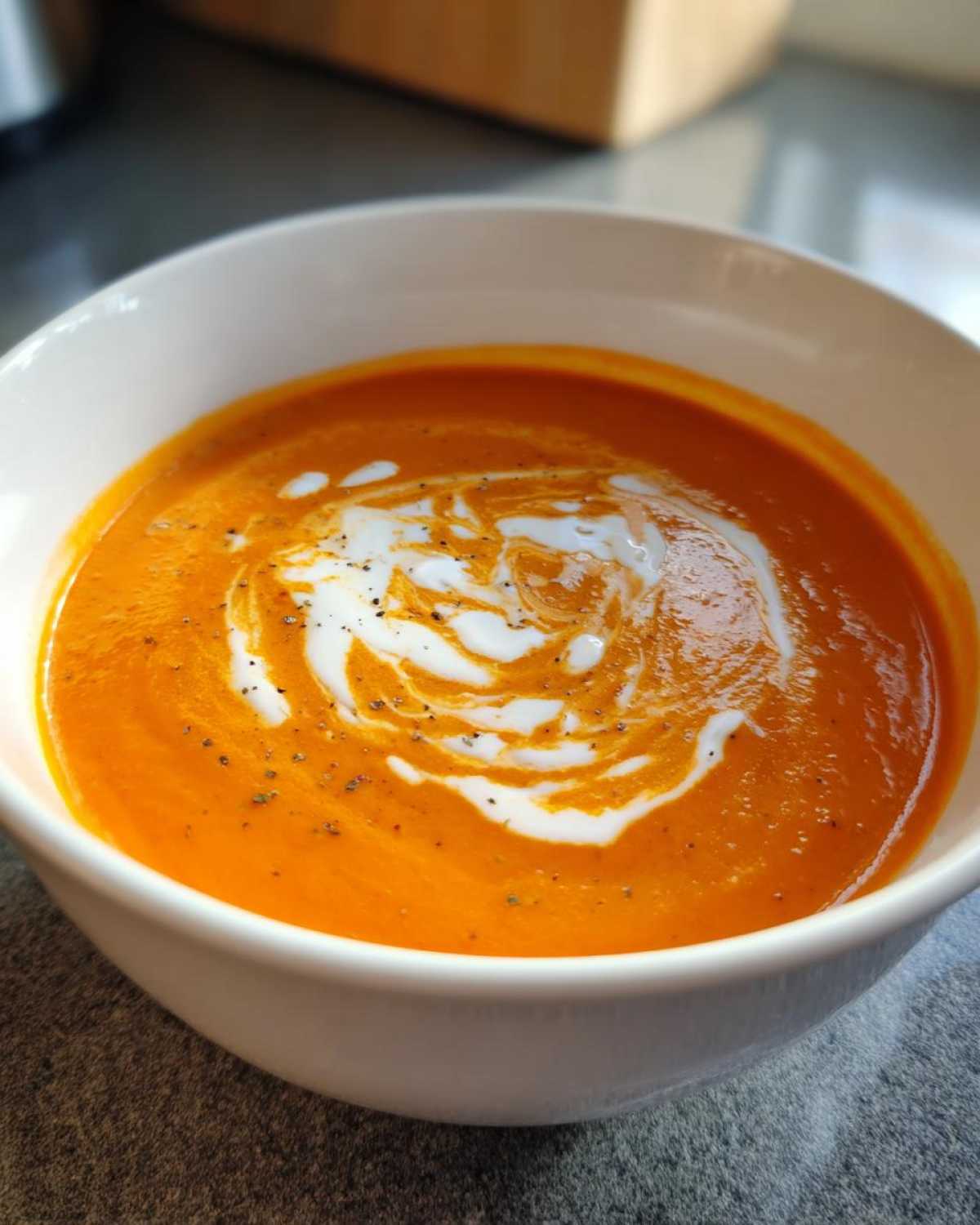 Close-up of a bowl of vibrant orange tomato bisque topped with a swirl of cream and cracked black pepper.